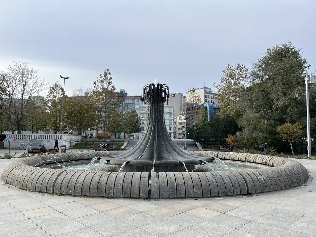 Gezi Parkı'nda taş havuzlu büyük fıskiye; ortada koyu renkli heykelsi fıskiye gövdesi, çevrede ağaçlar ve binalar.