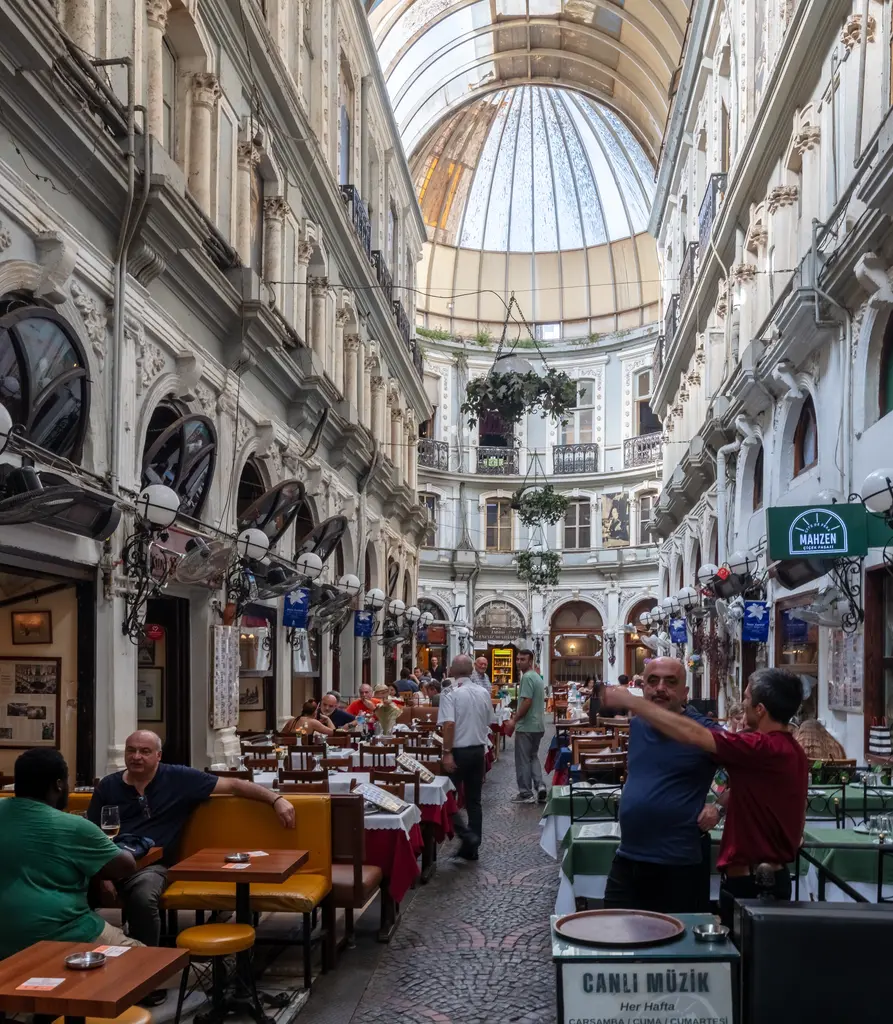 Çiçek Pasajı'nda meyhane masaları; geçit boyunca kurulmuş örtülü masalar, sohbet eden insanlar, sarkan çiçek sepetleri ve uzakta rotonda.