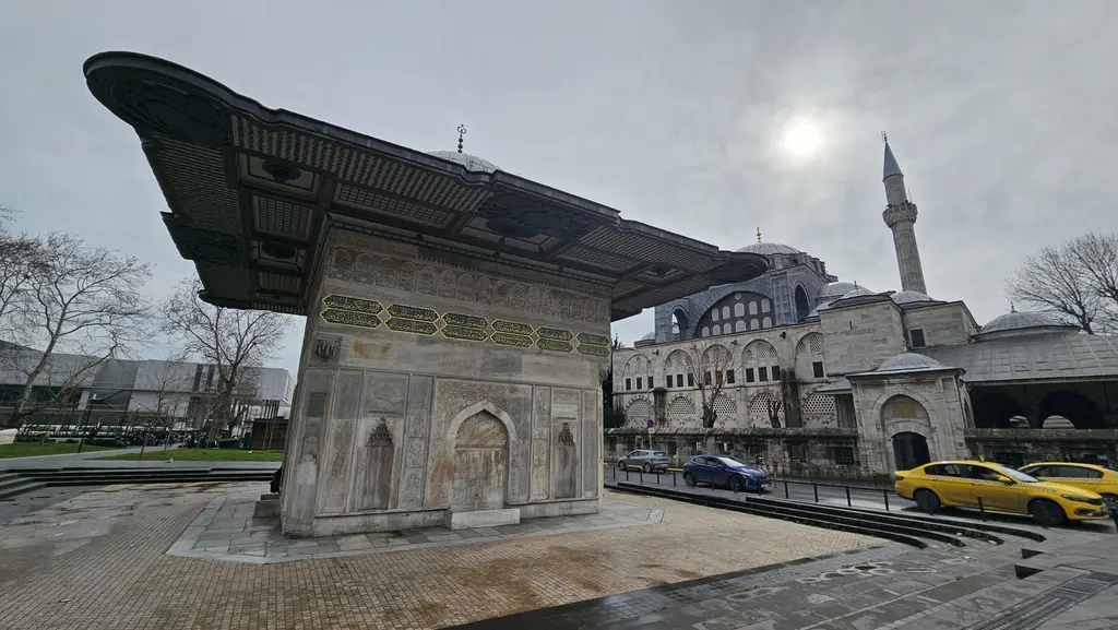 Kış gününde Tophane Çeşmesi; bulutlu gökyüzü, mermer gövde ve nişler, saçak ve arkada çıplak ağaçlar ile meydanın taş döşemesi.