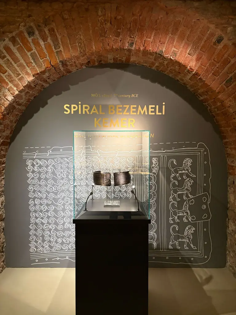 Spiral Bezemeli Kemer; tuğla kemerli nişte cam vitrin, gri duvarda sarmal desen çizimleri ve altın harflerle yazılmış başlık.