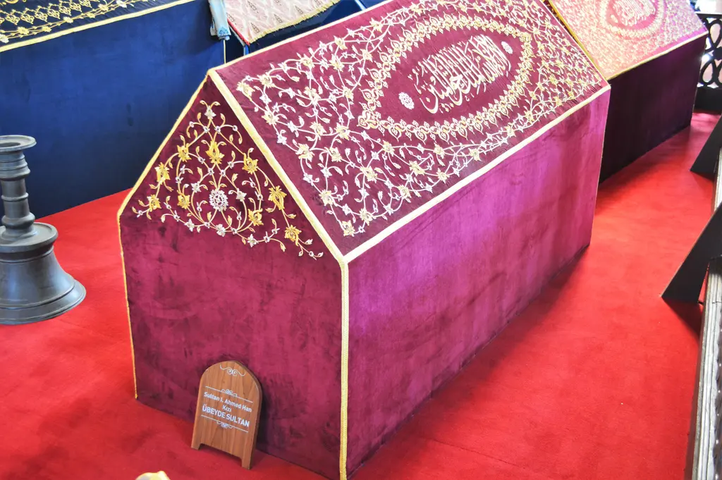 Übeyde Sultan'ın sandukası; kırmızı halı üstünde altın işlemeli mor kadife örtü, ortada yazılı madalyon ve ön yüzde ahşap isim levhası.
