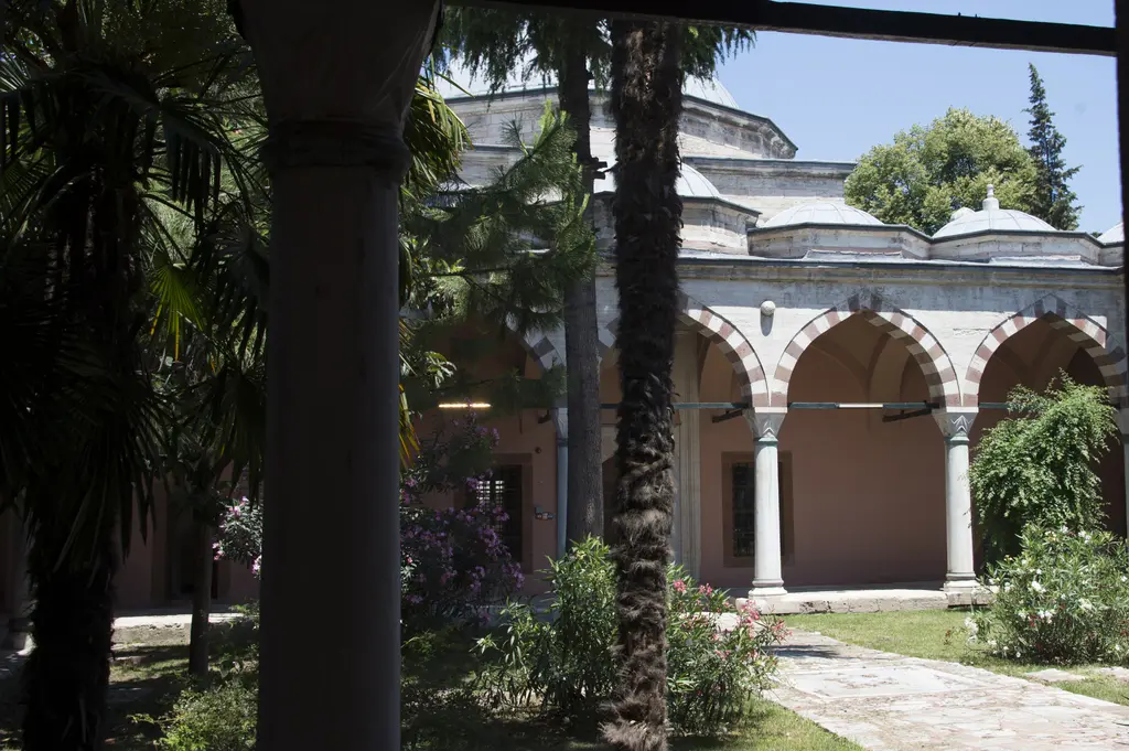 Haseki Sultan Külliyesi'nde palmiyeler arasından görünen revak; kırmızı ve beyaz taşlı kemerler, ince sütunlar ve bahçe.