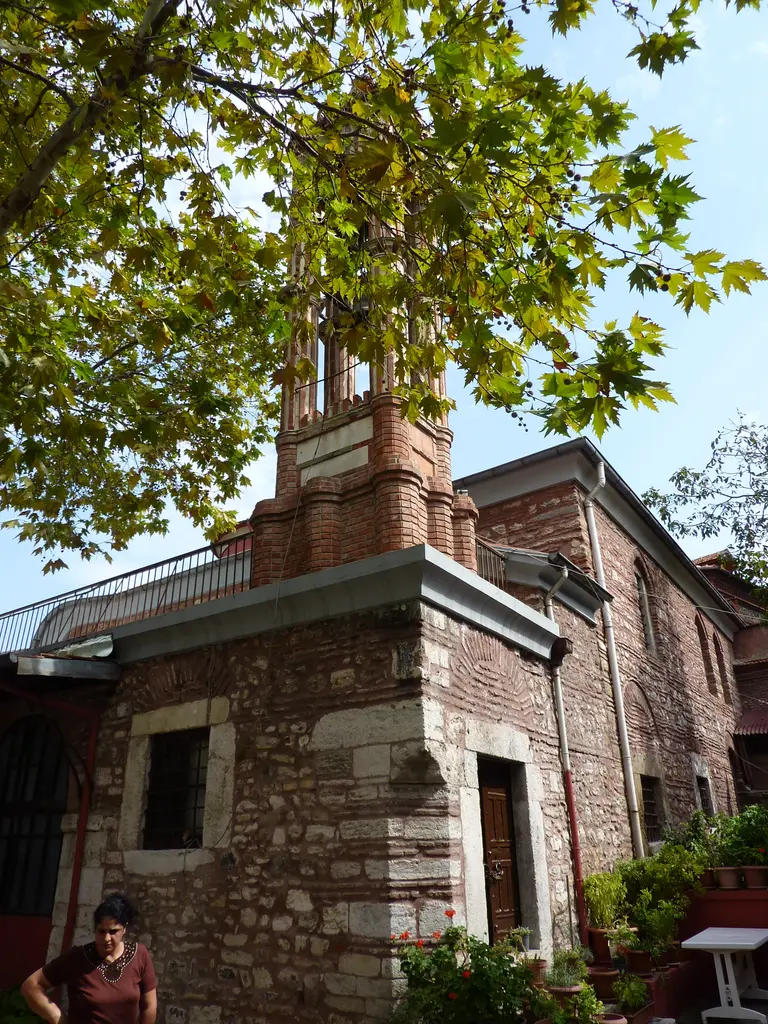 Kanlı Kilise'nin avlusunda çan kulesi; tuğla ve taş gövde, sivri tuğla çatı, önünde çınar dalları ve saksılarda çiçekler.