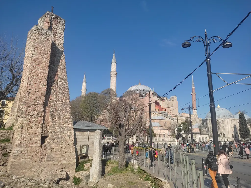 Milyon Taşı ile su terazisi; önde tuğla örgülü kule ve dibinde mermer ayak, arkada Ayasofya, tramvay direkleri ve kaldırımda yürüyenler.