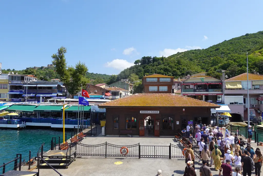 Anadolu Kavağı'nda iskeleden inen kalabalık; ahşap iskele binası, rıhtımdaki tur tekneleri ve arkada lokantalar.