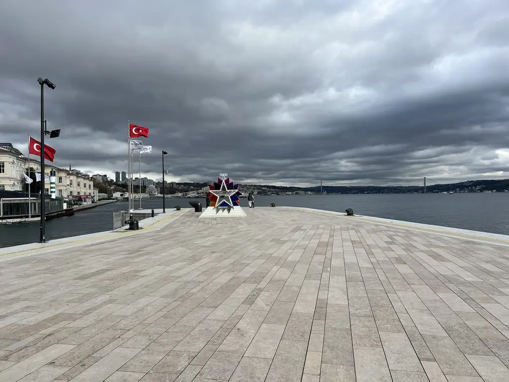Galataport'ta geniş taş iskele; ahşap desenli zemin, kenarda bayraklar ve heykel, arkada bulutlu gökyüzü altında Boğaz.