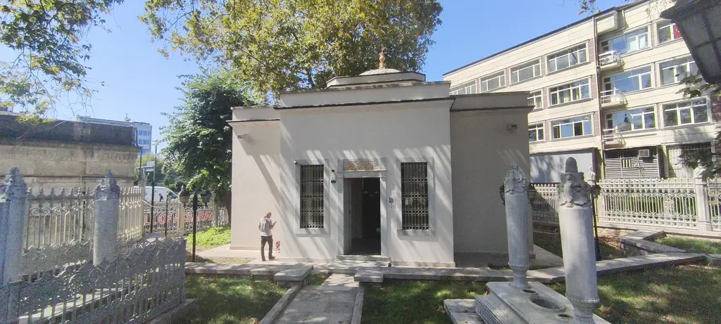 Pertevniyal Valide Sultan Türbesi; beyaz taş duvarlı küçük yapı, önünde çimen, mezar taşları, ağaçlar ve arkada çok katlı bina.