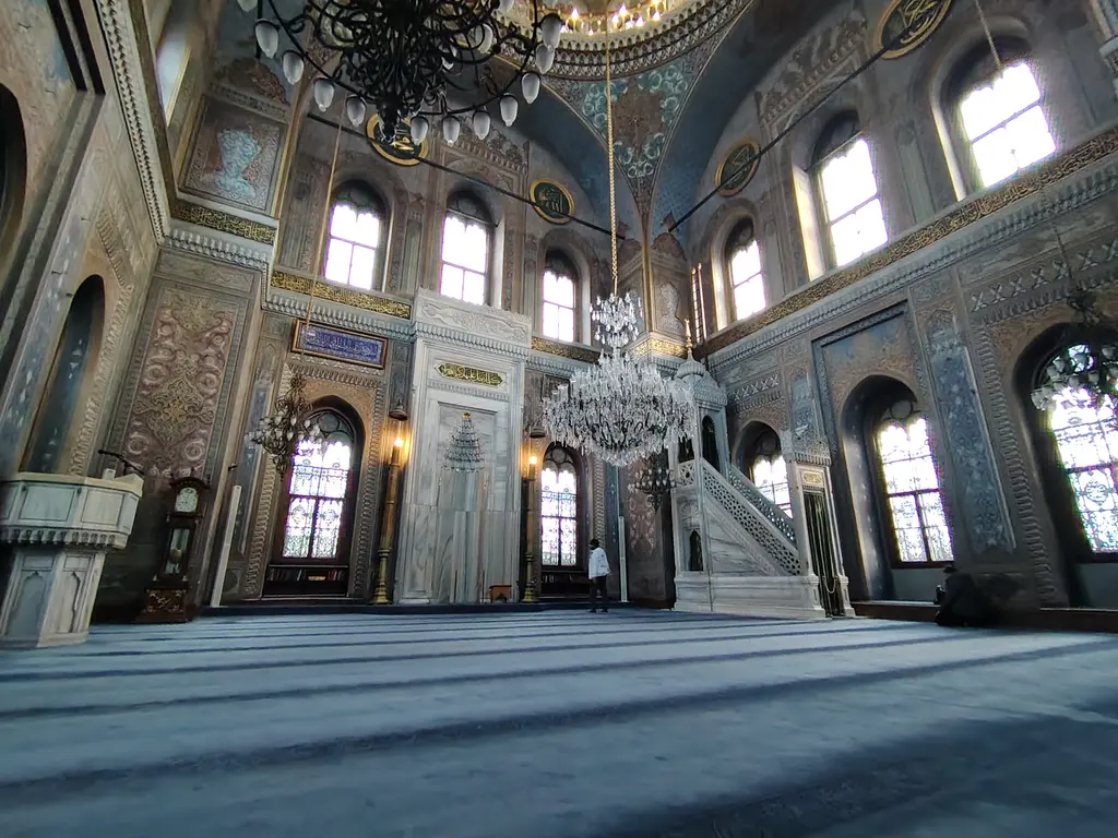Pertevniyal Valide Sultan Camii'nin harimi; mavi halı, mermer mihrap ve minber, kristal avize, sivri kemerli pencereler.