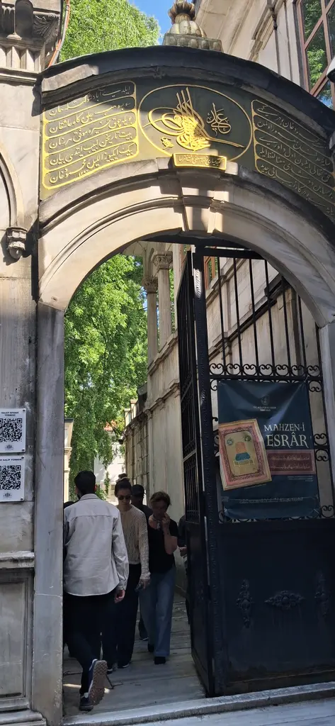 Galata Mevlevihanesi'nin kemerli taç kapısı; üstünde yaldızlı yazılı kitabe, açık demir kanatlar ve içeri giren ziyaretçiler.