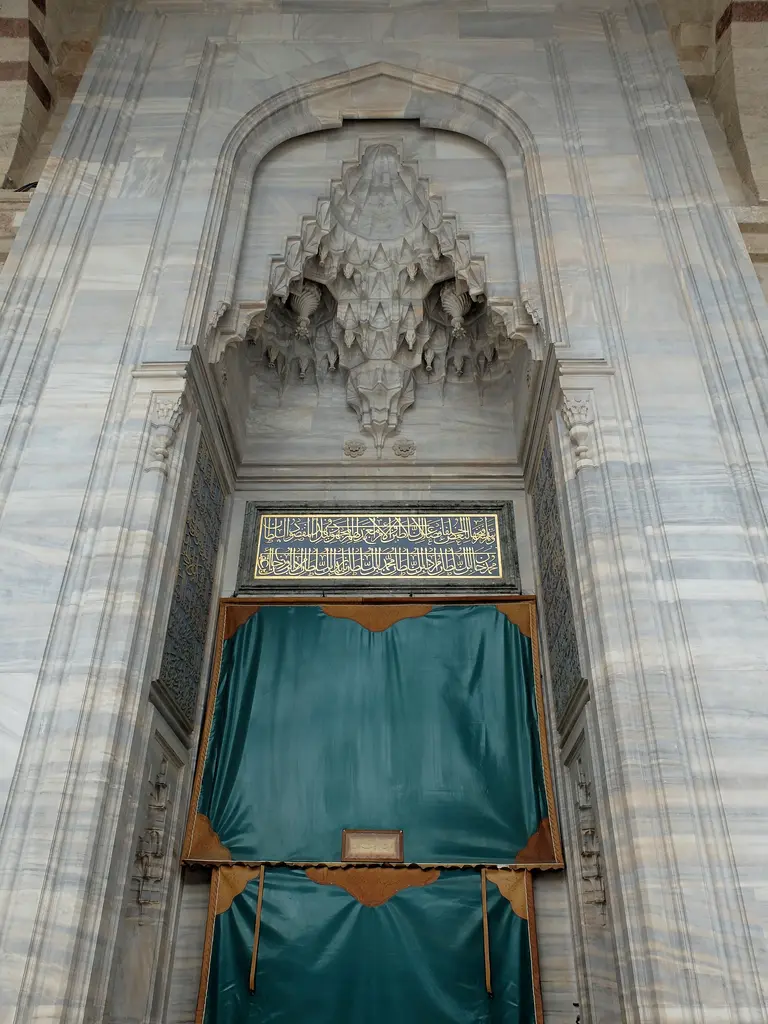 Fatih Camii'nde revaktan harime açılan kapı; yüksek gri beyaz mermer çerçeve, derin hücreli mukarnas kavsara, altın harfli kitabe ve açıklığı örten turkuaz yeşil perde.