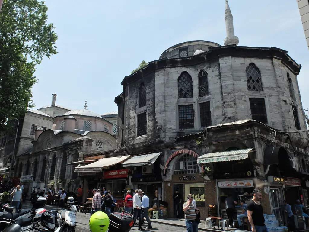 Nuruosmaniye Külliyesi'nin sokak köşesindeki yapısı; isle kararmış çokgen taş gövde, altında kafe ve büfe dükkânları, arkada minare şerefesi, önde motosikletler.