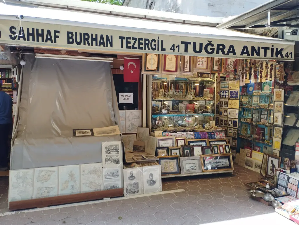 Sahaflar Çarşısı'nda bir antikacı cephesi; kırmızı tabela, ortada inik krem tente, iki yanda duvara dizilmiş eski harita, gravür ve portre çerçeveleri, altta çekmecelerde tasnif edilmiş baskılar.