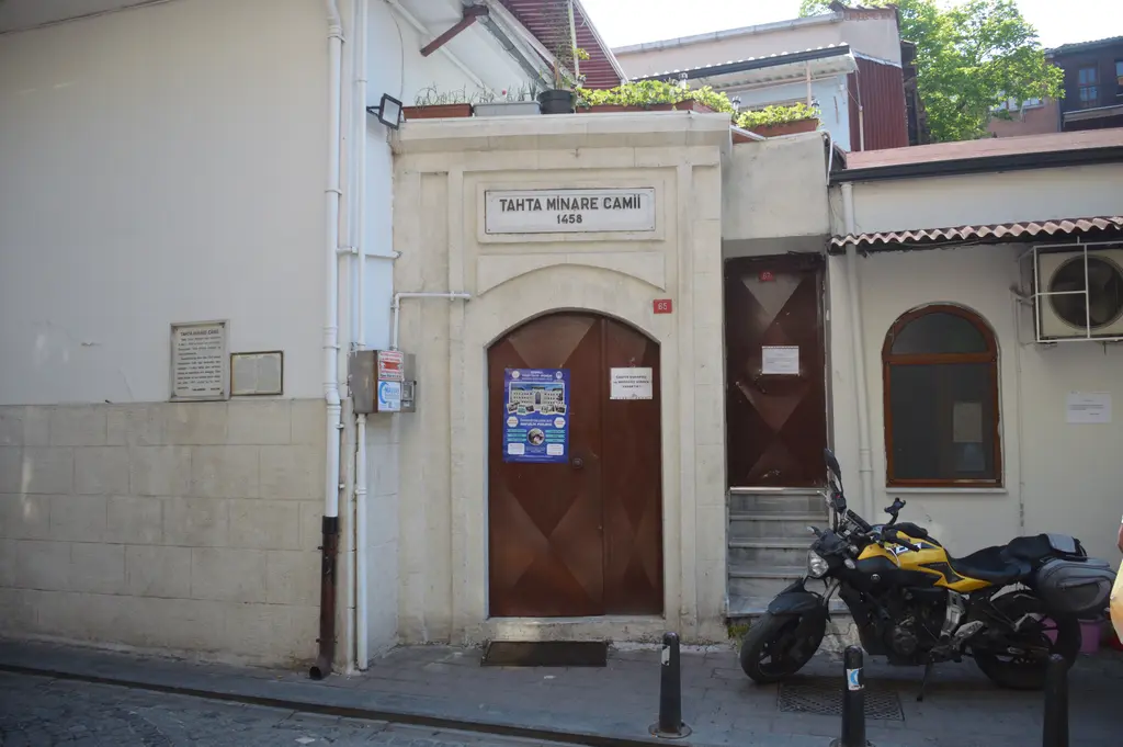 Tahta Minare Camii'nin girişi; krem cephe, kemerli ahşap kapı, üstte cami tabelası ve önde park etmiş motosiklet.