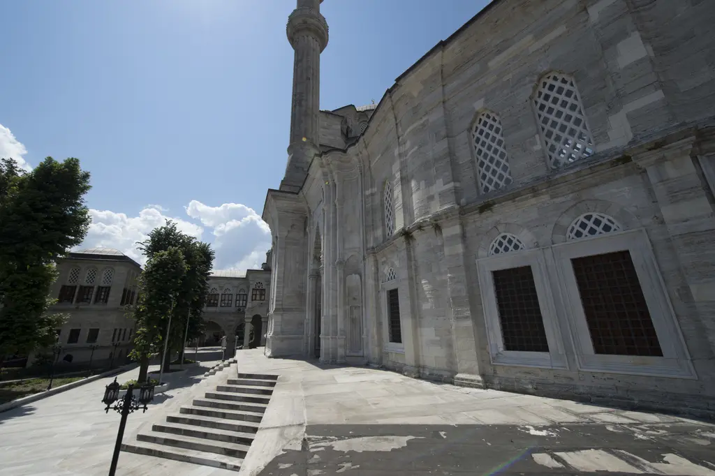 Nuruosmaniye Camii'nin avlusuna çıkan geniş taş merdiven; sağda minare gövdesi, üstte kemerli revak, iki yanda yapraklanmış ağaçlar ve açık gökyüzü.