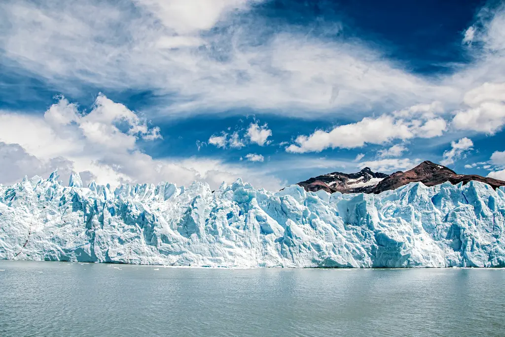 Arjantin'de Perito Moreno buzulunun göl suyuna inen buz cephesi; mavi-beyaz buz kuleleri, arkada karlı dağlar ve bulutlu mavi gökyüzü.