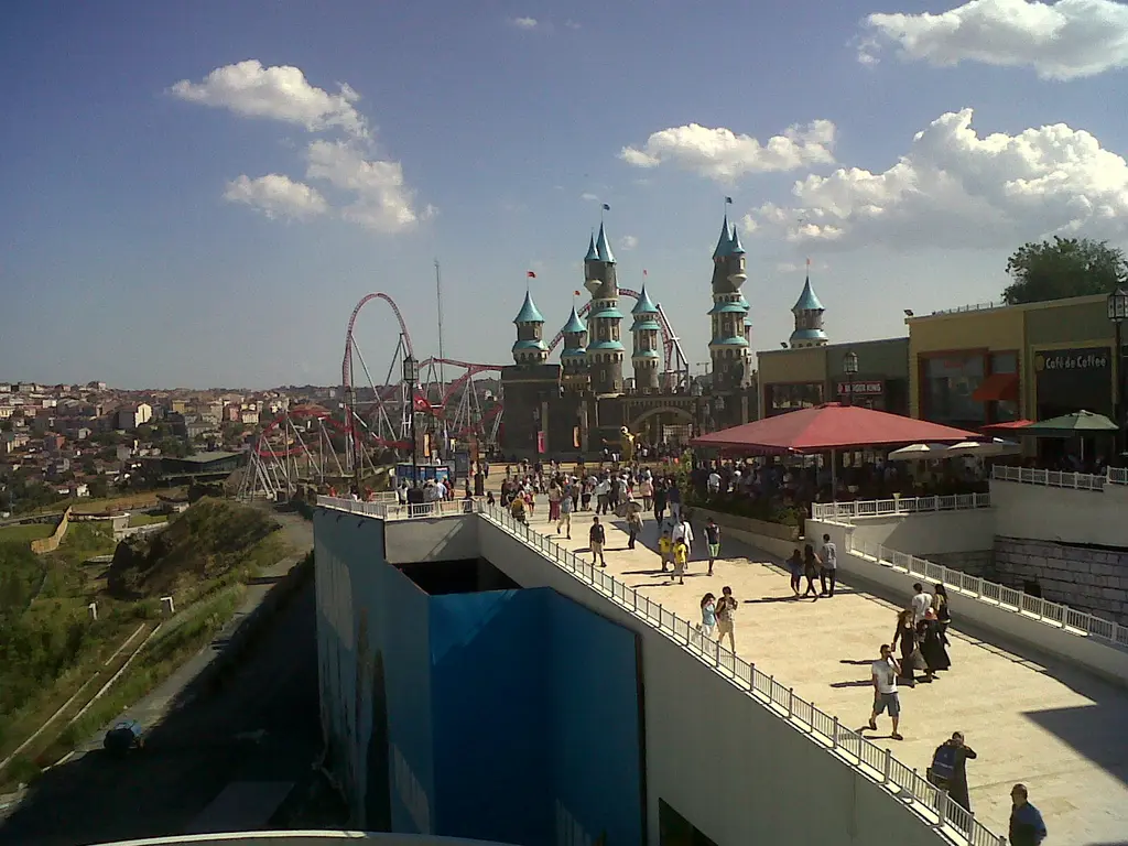 Vialand'de alışveriş caddelerinden parka uzanan geniş yürüyüş yolu; ilerde şato kuleleri ve roller coaster, yanlarda kafeler ve ziyaretçiler.