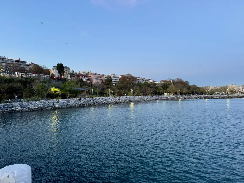 Moda kıyısı; dalgakıran kayalar, sakin deniz, yamaçta ağaçlar ve binalar, akşam ışığı.