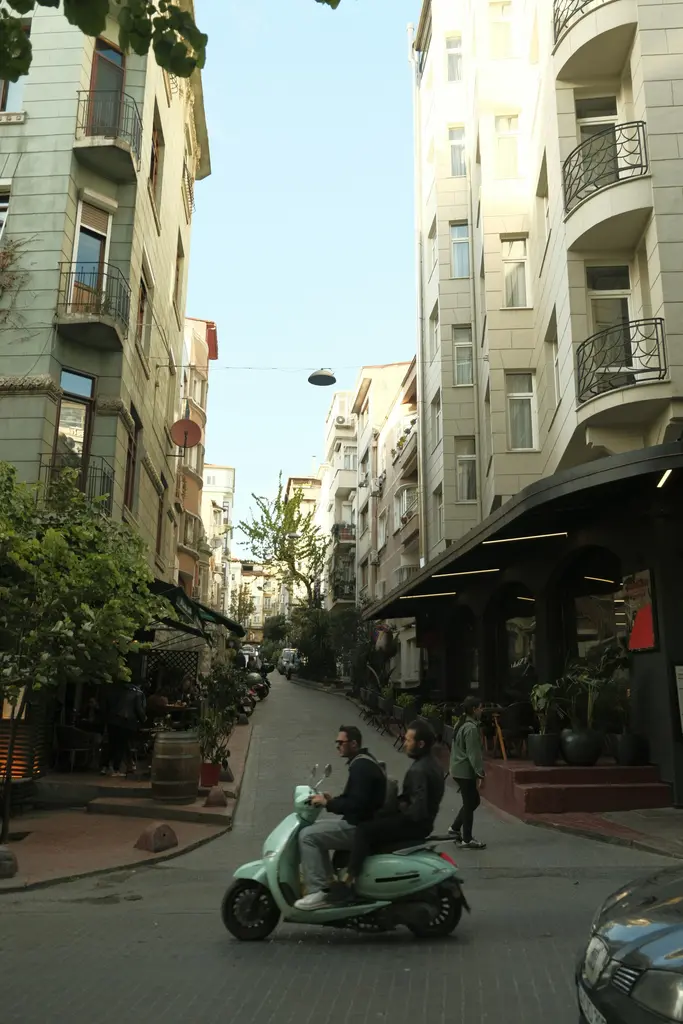 Cihangir'de ağaçlı bir sokak; iki yanda balkonlu apartmanlar, köşede bir kafe ve yokuşu inen motosikletli iki kişi.