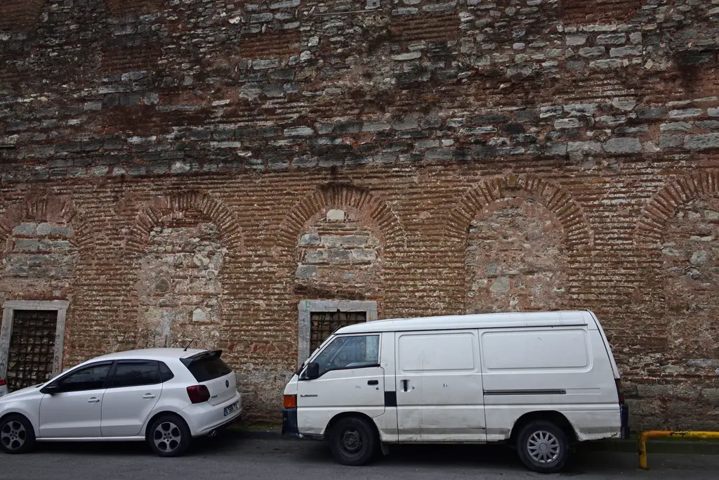 İmrahor İlyas Bey Camii'nin sokağa bakan duvarı; almaşık tuğla ve taş örgüde sıralanan kör kemerler, altta örülmüş kapı boşlukları, önde park etmiş beyaz araçlar.