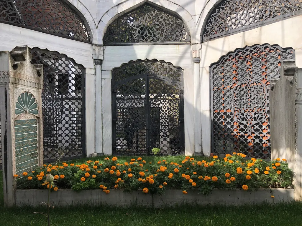 Yeni Valide Camii'nde türbe; bronz şebekeli kemerli açıklıklar, mermer sütunlar ve önünde turuncu çiçekler.