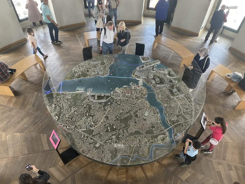 Galata Kulesi'nde yuvarlak bir İstanbul maketi; mavi Boğaz ve Haliç, beyaz yapı dokusu ve maketin çevresinde gezen ziyaretçiler.