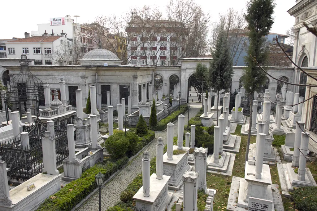 II. Mahmud Türbesi hazireye genel bakış; sıra sıra mermer mezar taşları, taş döşeli yol, yeşil çitler, arkada kemerli revak ve kubbeli yapı.