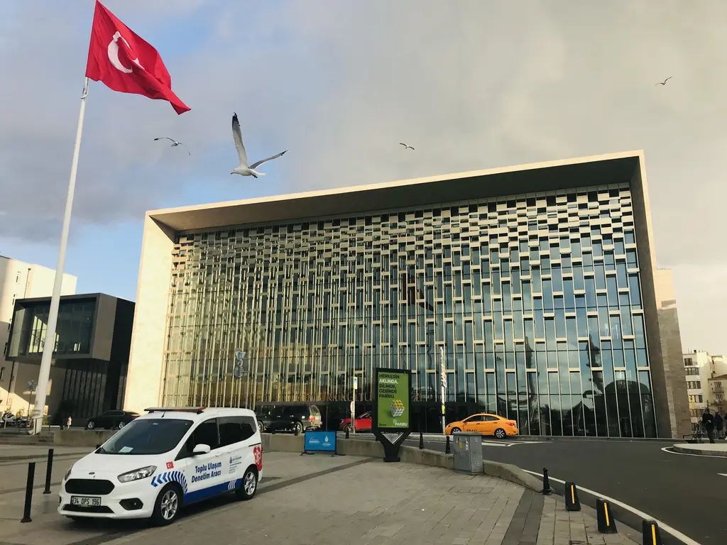 Atatürk Kültür Merkezi yandan; taş kaplı yan duvar, cam cephe, önde Türk bayrağı, uçan martılar ve park etmiş bir araç.