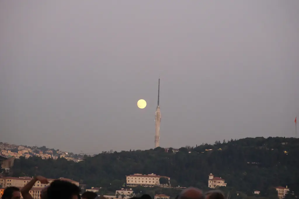 Alacakaranlıkta Çamlıca Kulesi'nin silueti; kulenin yanında yükselen dolunay, ağaçlık tepe ve önde izleyicilerin başları.
