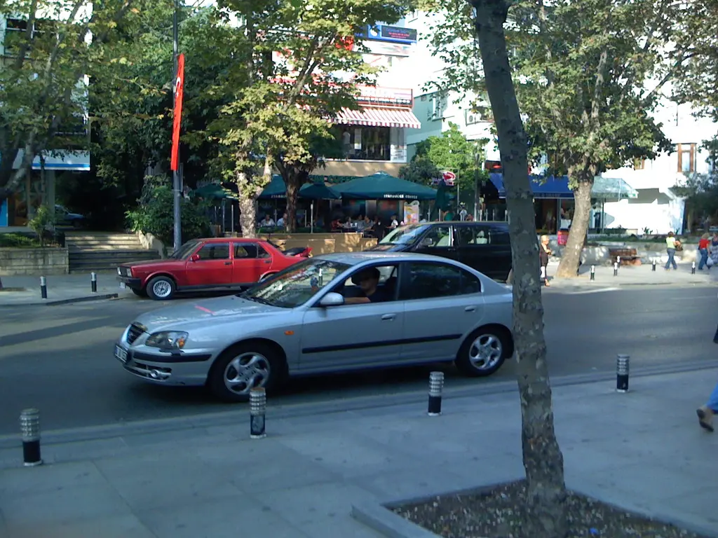 Suadiye'de Bağdat Caddesi; ağaçların altında park etmiş arabalar, kırmızı eski otomobil, kafe tenteleri ve bayraklar.