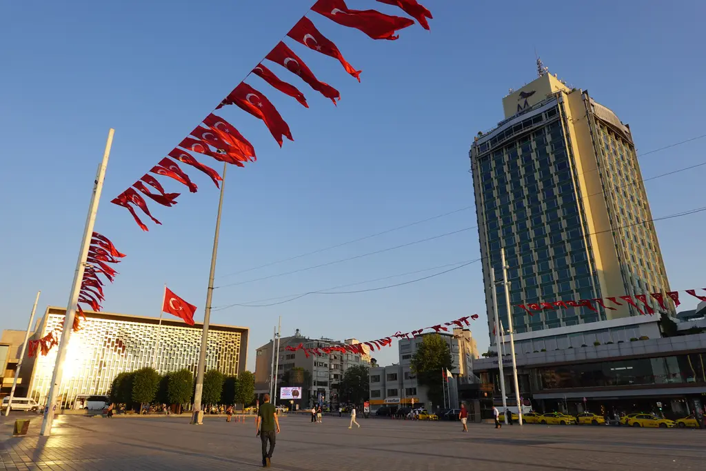 Güneşli bir akşamüstü Taksim Meydanı; gökyüzüne gerilmiş Türk bayrakları, sağda yüksek otel binası, solda Atatürk Kültür Merkezi'nin cephesi.