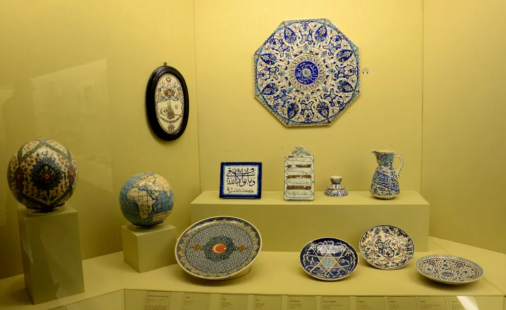 Sarı duvarlı bir vitrinde Kütahya çinileri ve seramikleri; mavi desenli tabaklar, sekizgen bir pano, sürahi ve kâseler.