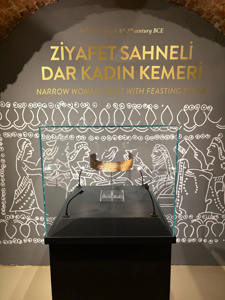 Ziyafet Sahneli Dar Kadın Kemeri; cam vitrinde kavisli bronz kemer, arkada ziyafet ve figür çizimleri ve iki dilli altın başlık.