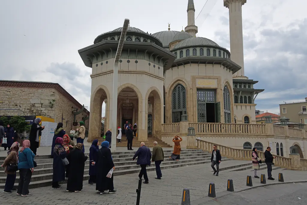 Taksim Camii'nin girişi; geniş taş basamaklar, kemerli kapı açıklıkları, küçük kubbeli köşe kuleleri ve merdivende yürüyen ziyaretçiler.