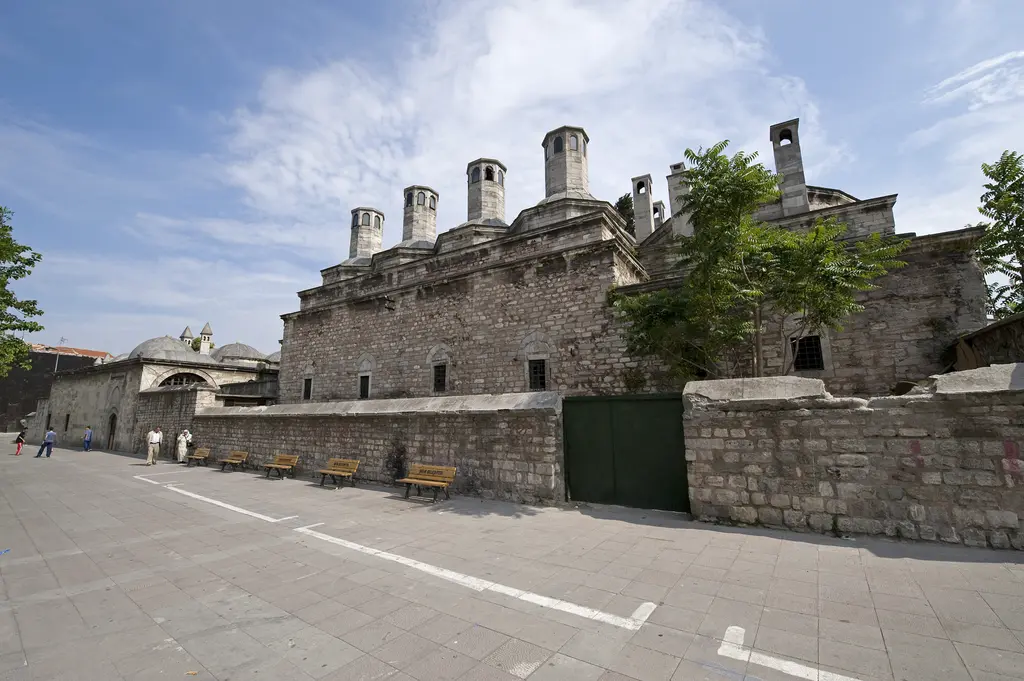 Haseki Sultan Külliyesi'nin cadde cephesi; alçak taş duvar, arkada kubbeler ve bacalar, yeşil bir kapı, ağaçlar ve taş döşeli meydan.