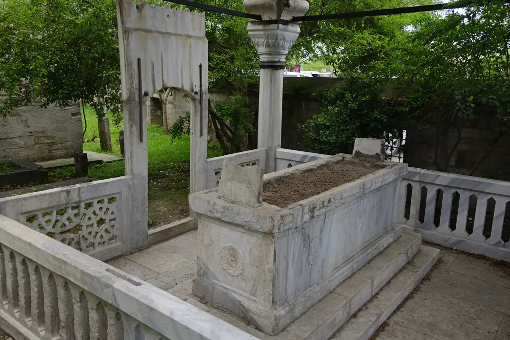 Defterdar Mahmud Çelebi'nin sandukası; mermer korkuluklarla çevrili taş sanduka, köşe sütunu, ağaçlar ve hazire.