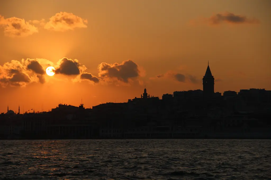 Gün batımında Karaköy ve Galata Kulesi'nin silueti; turuncu gökyüzü, bulutların arasında güneş ve karanlık su.