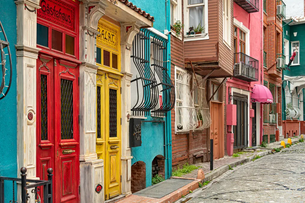 Balat'ta kırmızı ve sarı boyalı iki ahşap kapı; mavi ve pembe cepheler, demir parmaklıklı pencereler ve yokuş sokak.