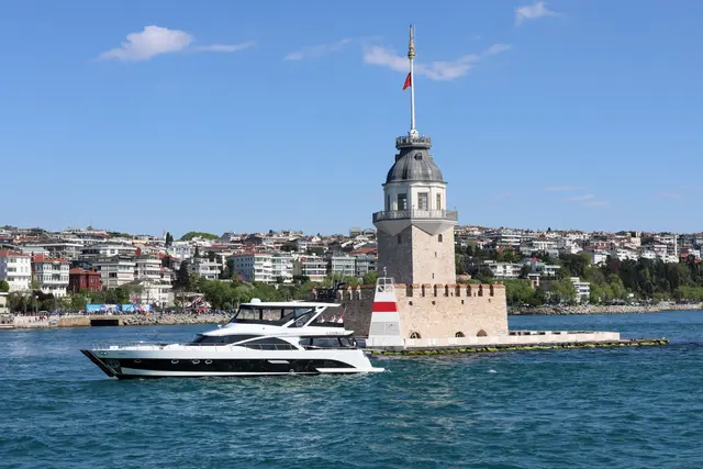 Üsküdar açığında Kız Kulesi; taş kaide üzerinde beyaz kule, tepesinde bayrak, önünden geçen yat, arkada Üsküdar'ın evleri.
