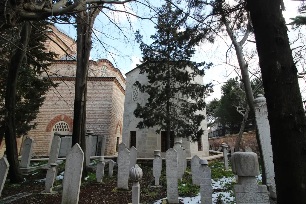 Rum Mehmed Paşa Camii'nin haziresi; ağaçlar arasında eski mezar taşları, arkada türbe ve caminin tuğla duvarı.