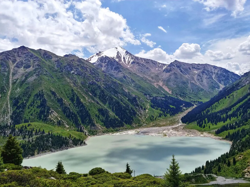 Büyük Almatı Gölü; dağların arasına sıkışmış turkuaz su yüzeyi, kıyısındaki ladin ormanı ve arkada sıralanan karlı zirveler.
