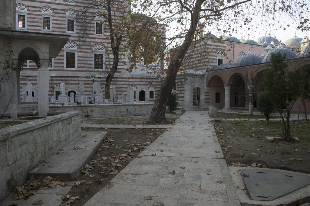 Zal Mahmud Paşa Külliyesi'nin alt bahçesi; taş döşeli yol, ağaçlar, mezarlar, sağda kubbeli medrese revakları ve arkada caminin duvarı.