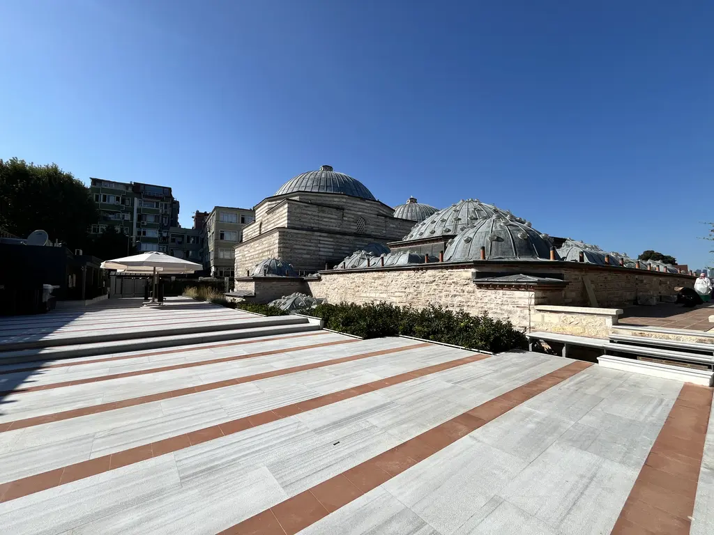 Çinili Hamam'ın terası; kahverengi gri şeritli mermer döşeme, gerisinde hamamın kubbeleri ve taş duvarı, şemsiye, ağaçlar ve apartmanlar.