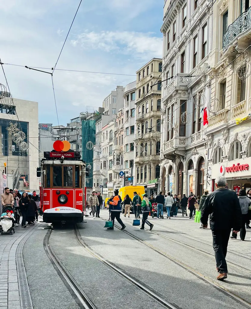 İstiklal Caddesi'nde kırmızı tramvay; arkada balkonlu ve süslü taş cepheli apartmanlar, üstte tramvay telleri ve bulutlu gökyüzü.