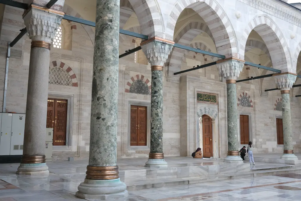 Avlunun revak altından bakış; devşirme sütunlar, sivri kemerler, mermer döşeme ve karşıda caminin kemerli kapı ile pencere sırası.