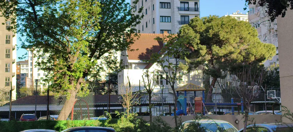Göztepe'de Atlı Muazzez Hanım Köşkü; ağaçların arasında kırmızı çatılı beyaz yapı, önünde oyun alanı ve apartmanlar.