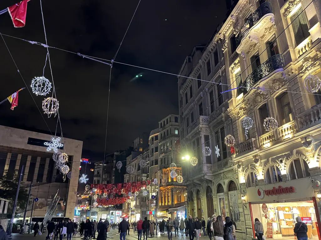 Gece İstiklal Caddesi'nde ışıklandırılmış süslü taş cepheler; caddeye asılı ışıklı toplar ve aşağıda yürüyen kalabalık.