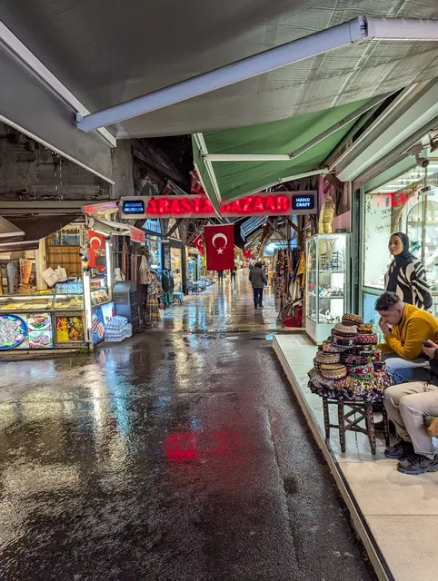 Arasta Çarşısı'nda ıslak mermer zeminli sokak; iki yanda dükkânlar, yukarıda yeşil tenteler, asılı Türk bayrakları ve tabure üzerinde oturan bir adam.