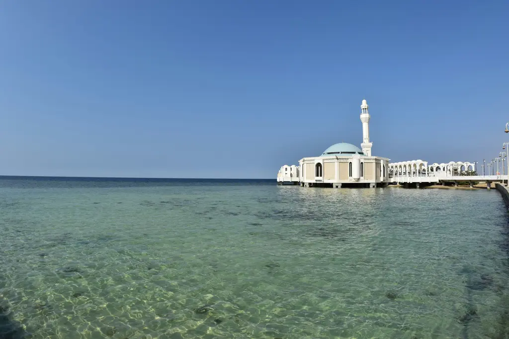 Cidde'de deniz üzerinde yükselen beyaz cami; kubbesi, tek minaresi, kıyıya bağlanan platformu ve önündeki berrak turkuaz su.