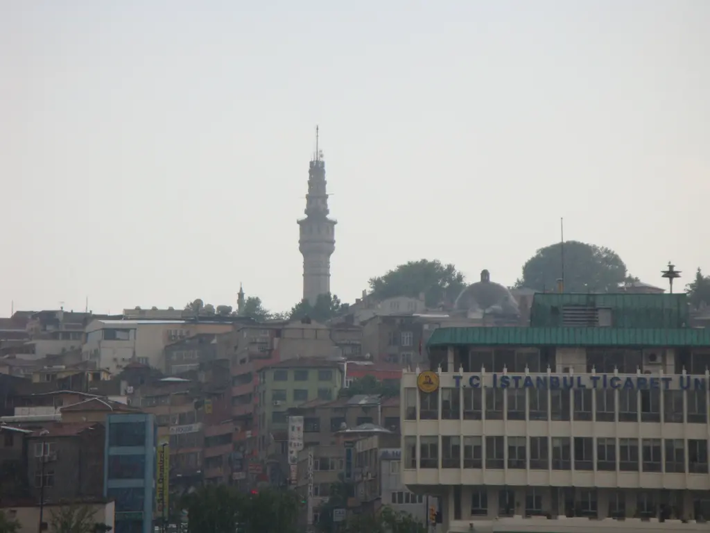 Uzaktan Beyazıt Kulesi; şehir çatılarının üstünde yükselen ince gövde, çevresindeki apartmanlar ve puslu gökyüzü.