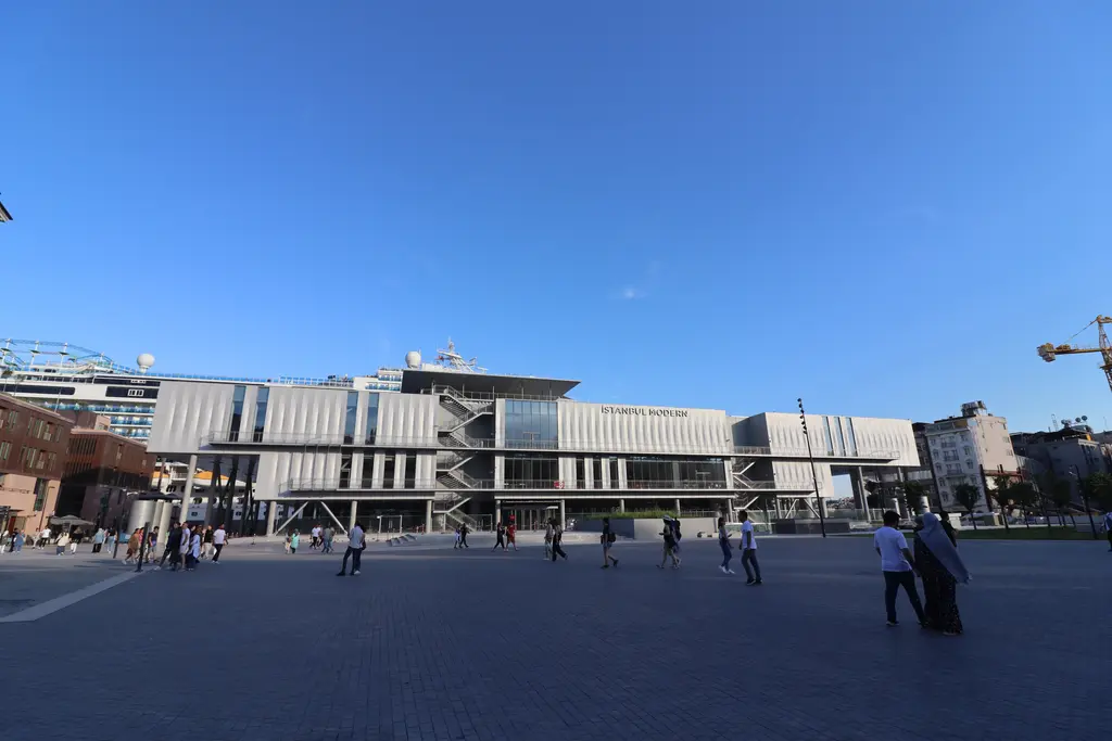 Mavi gökyüzü altında İstanbul Modern'in uzun gri cephesi; önde taş döşeli meydan ve yeşil bir çimlik şerit.